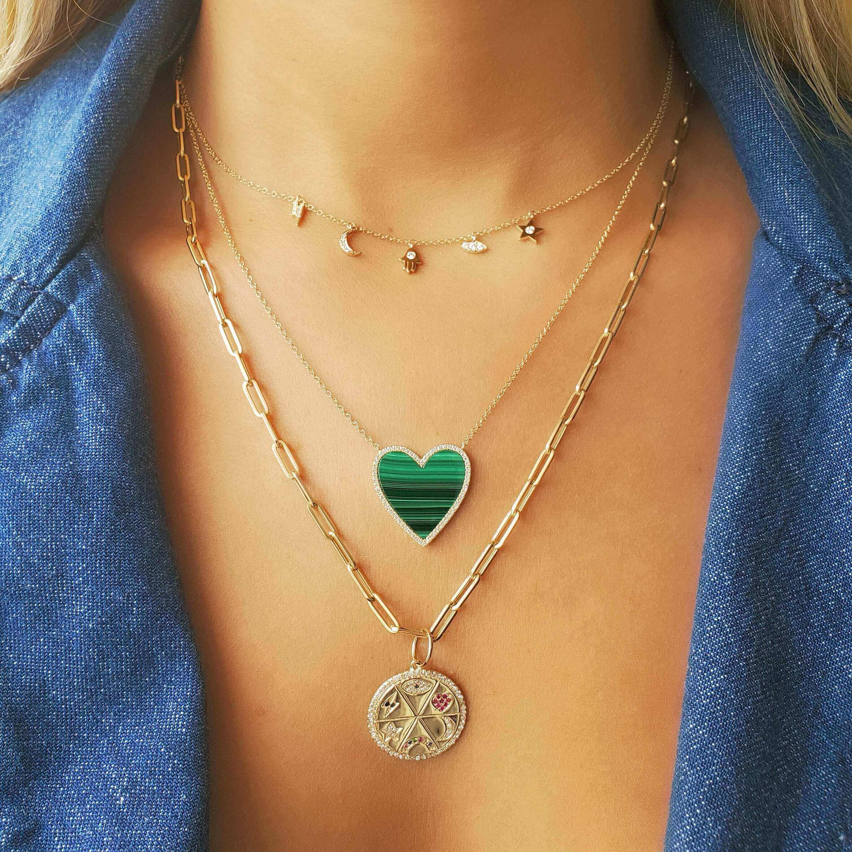 Woman wearing layered gold necklaces with a heart-shaped pendant, celestial charms, and a coin medallion, styled with a denim shirt