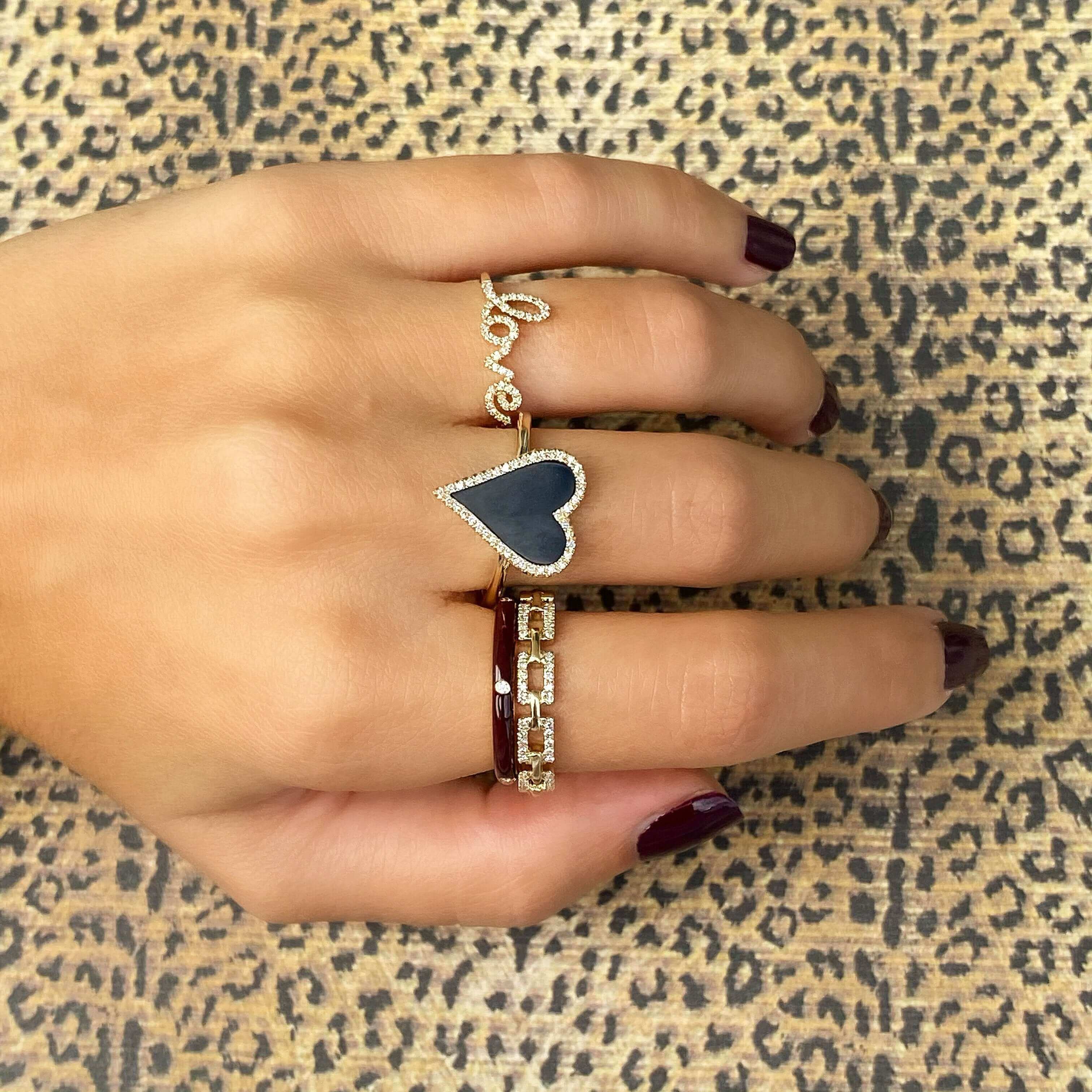 Elegant hand wearing gold jewelry with heart-shaped black stone ring and love script ring, set against a leopard print background.
