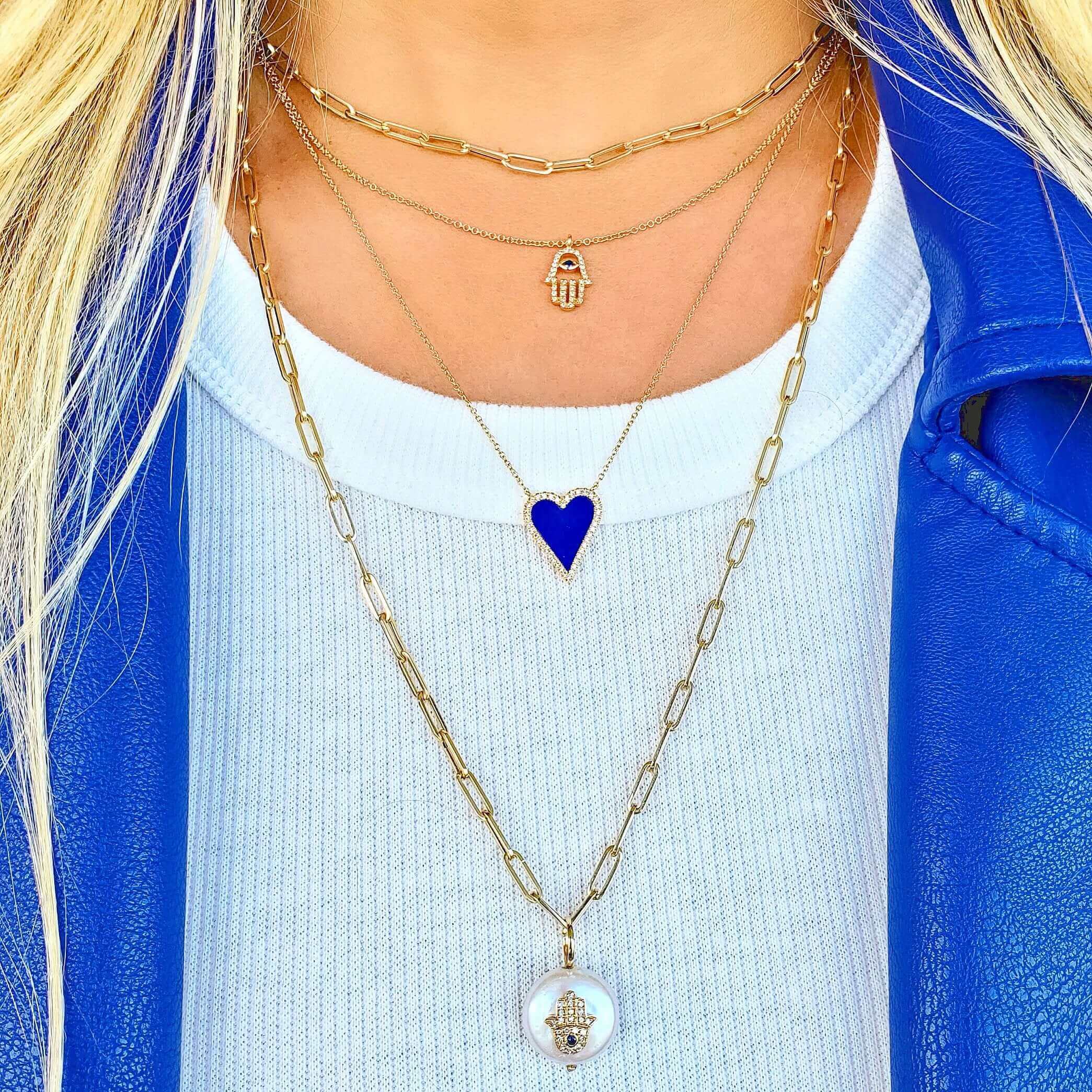 Layered gold necklaces with a Hamsa charm, blue heart, and pendant, styled with a blue jacket and white shirt, showcasing luxury jewelry brand.